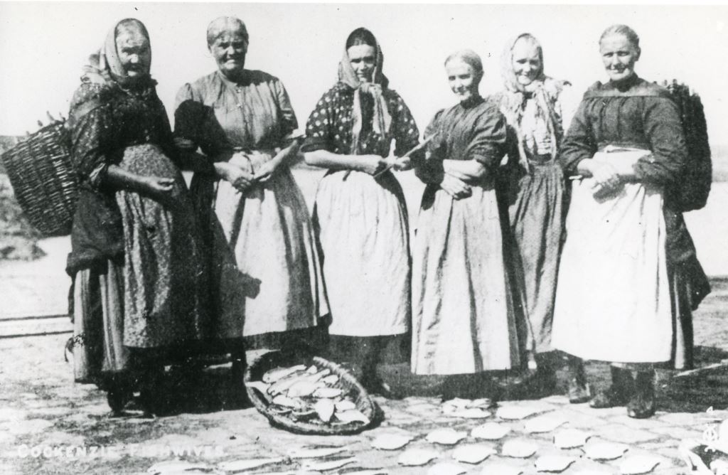 A group of six women dressed in traditional fishwives' clothing with long aprons. Fish are laid out on the ground in front of them, some in a basket. Two of the women carry creels on their backs. Image dates to September 1910.