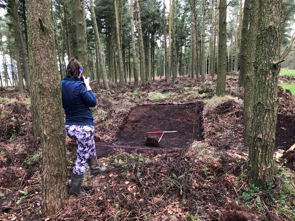 Image of a female archaeologist in forestry, taking a photograph of a ditch which has been cleared of vegetation where a trench has been laid out