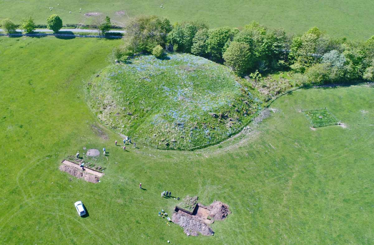 Aerial photograph showing a round, grass-covered mound on which the remains of the castle sit, with trenches open in the flat grassy field in front of the motte