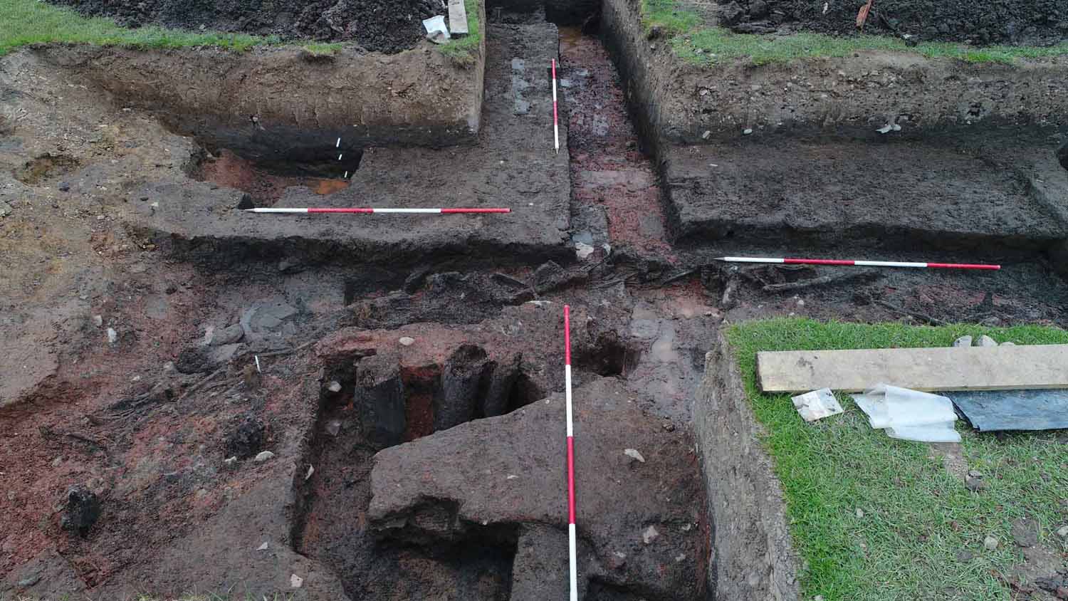 Photograph of a cross-shaped trench with a curving feature made of posts and wattle running diagonally across the centre, and some much larger posts in front of these