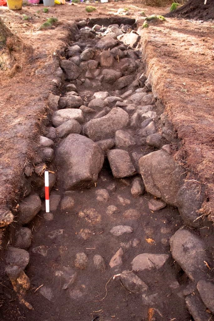 A long trench with stones forming the body of the cairn in the background, and a stony, possible trampled ground surface in the foreground