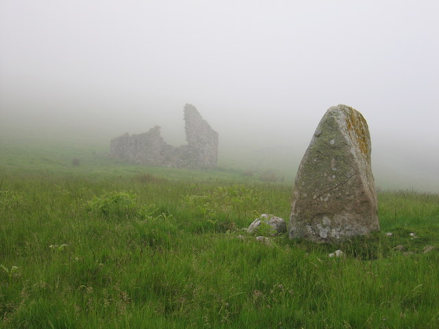 A standing stone in the foreground, with a ruinous building in the mist behind