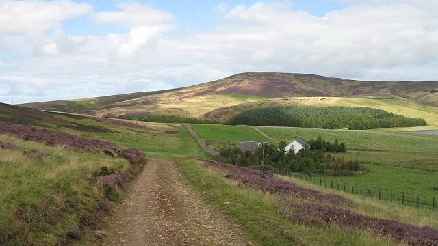 Rough track across open landscape, with two houses to the right of the track and heather-covered hills in the distance