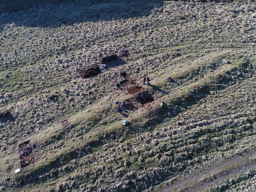 Aerial photo showing the outline of a long, grass-covered feature with trenches over it and people working